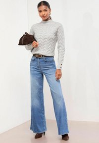 Jasnoszary sweter w splot kabelkowy, wysoka szyja, długie rękawy; jasnoniebieskie szerokie dżinsy; brązowa zamszowa torba; brązowe botki na obcasie.