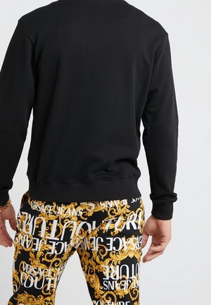 Mann trägt schwarzes Langarmshirt und schwarze Hose mit goldenem Barockmuster und weißem Versace Jeans Couture-Schriftzug, Rücken zur Kamera.