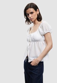 Kvinde med mørkt hår iført guldøreringe, en hvid tekstureret bluse med pufærmer, der er knyttet foran, og mørkeblå jeans med den ene hånd i lommen.