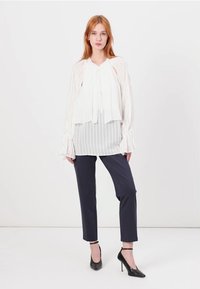 Blusa bianca con righe verticali, con lacci al collo, abbinata a pantaloni slim blu scuro e scarpe con tacco nere. Tessuto liscio e leggero.