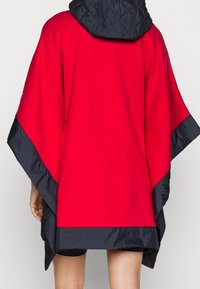 Röd poncho med en svart vadderad huva, med en bred, flödande design. Sömmen är avslutad i svart, vilket framhäver kontrasterande texturer.