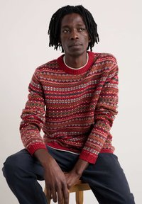 Sarkans džemperis ar zaļiem, zilajiem un baltajiem akcentiem. Biezs adījums un ribotās piedurknes. Saskaņots ar tumšiem biksēm, apsēdīsies uz koka krēsla.