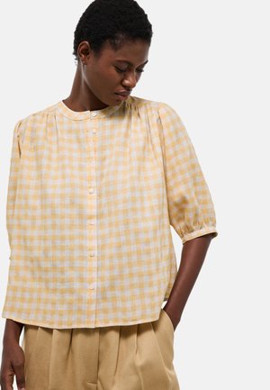 Femme portant un chemisier à carreaux jaune et blanc à boutons avec manches trois-quarts et pantalon plissé beige, regardant vers le bas sur le côté.