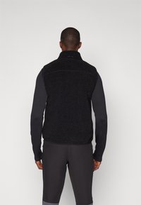 Μαύρο γιλέκο από φλις με ψηλό γιακά, υφή υφάσματος και κομψό σχέδιο. Φοριέται πάνω από ένα εφαρμοστό μπλουζάκι με μακρύ μανίκι και αθλητικά παντελόνια.