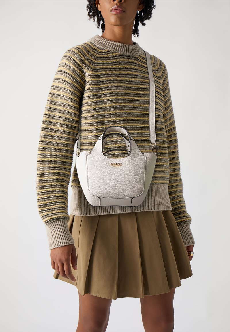 Pequeño bolso de cuero color crema con un acabado texturizado, forma redondeada, asas cortas y una correa de hombro desmontable. Llevado con un suéter a rayas y una falda.