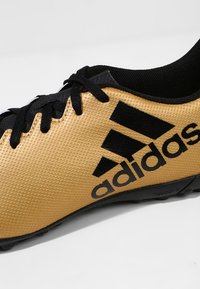 Kultaiset ja mustat Adidas-jalkapallokengät, joissa on teksturoitu pinta, mustat nauhat ja huomattava logo, suunniteltu suorituskykyä varten kentällä.