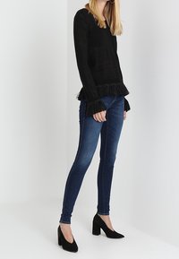 Černý svetr s texturovaným vzorem a třásněmi na spodním lemu, kombinovaný s tmavě modrými úzkými džíny a černými vysokými podpatky.