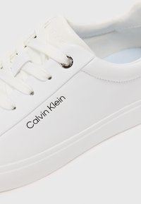 Bílé kožené tenisky s kulatou špičkou, stříbrným okem, textura šněrování a černým potiskem loga "Calvin Klein" na boku.