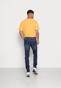 T-shirt de algodão amarelo, jeans azul-escuro com um patch de couro, e ténis brancos com detalhe de estrela preta, vistos de costas.