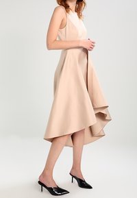 Beige ärmlös klänning med asymmetrisk nederkant och slät textur, i kombination med svarta lackläder högklackade mules.