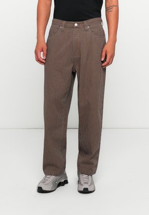 Man wearing brown vertical striped pants, black shirt, and gray Nike sneakers, standing against a plain white background.