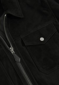 Melna zamšāda jaka ar detaļām, kas ietver rāvējslēdzēju, krūšu kabatu ar sprādzes aizdari un kontrasta šuvēm. Gluda tekstūra un pielāgota dizaina izpildījums.