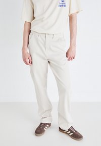 Pantalones de algodón beige con un diseño de pierna recta, que presentan lazos para cinturón, bolsillos laterales y una textura suave. Llevados con zapatillas deportivas marrones.