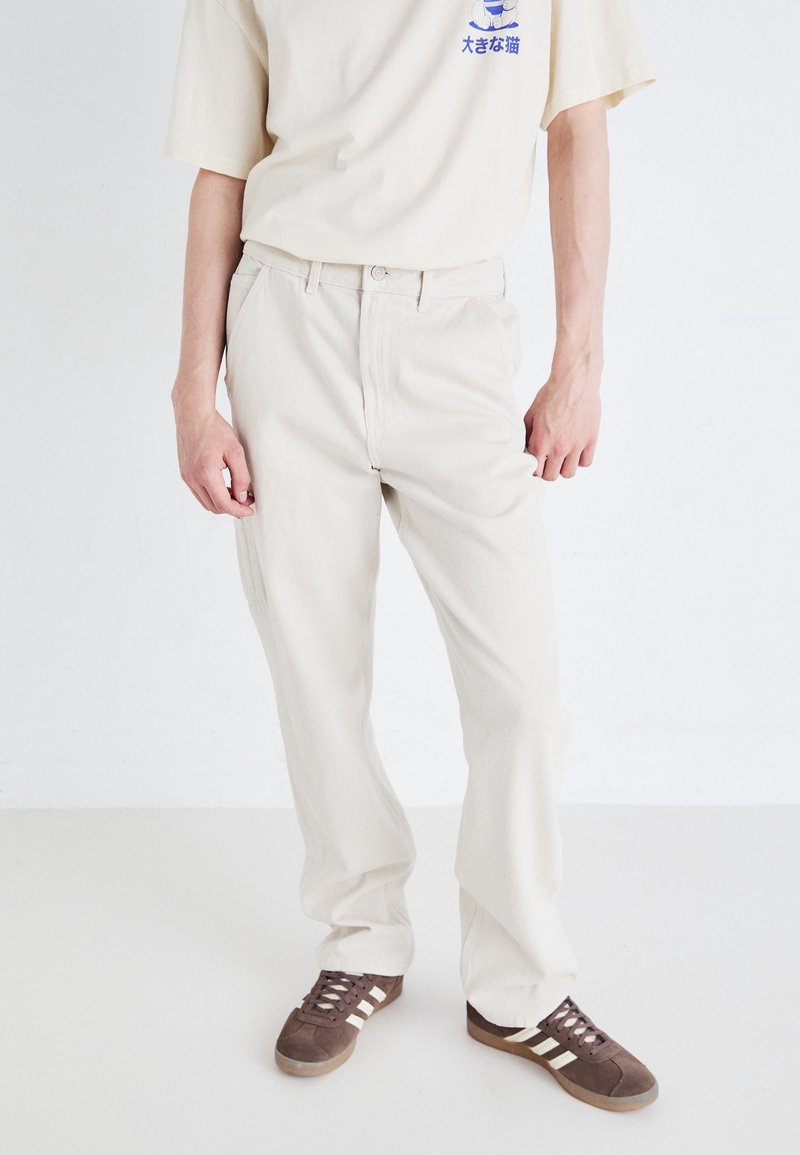 Pantalones de algodón beige con un diseño de pierna recta, que presentan lazos para cinturón, bolsillos laterales y una textura suave. Llevados con zapatillas deportivas marrones.