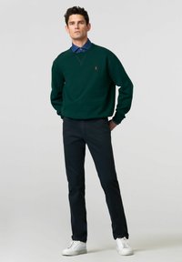 Donkergroene sweatshirt met een ronde hals en geborduurd logo, gecombineerd met zwarte broek en witte sneakers. Glad materiaal, ontspannen pasvorm.