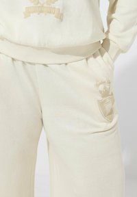 Kremowy dres charakteryzujący się miękką teksturą, elastycznym pasem i subtelnie haftowanymi detalami logo na klatce piersiowej oraz kieszeni.