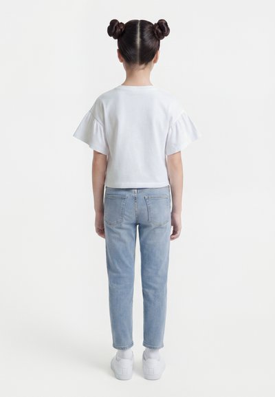 Νεαρή κοπέλα που στέκεται με την πλάτη, φορώντας λευκό πουκάμισο με κοντά μανίκια και βολάν, ανοιχτό μπλε τζιν, λευκές κάλτσες και λευκά αθλητικά παπούτσια, με τα μαλλιά της σε δύο κότσους.