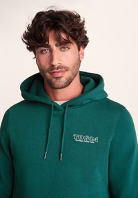 Green hoodie with drawstring, featuring a white logo on the left chest. Fabric appears soft with a smooth texture.