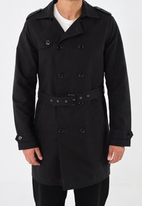 Svart dubbelknäppt trenchcoat med bälte i midjan, med knappdetaljer och sidofickor. Tillverkad av ett slätt, hållbart tyg.