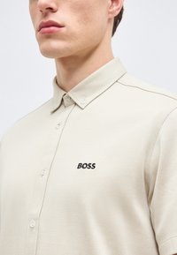 Gros plan sur une personne portant une chemise beige à col avec "BOSS" brodé en noir sur la poitrine.