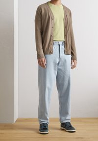 Hnědý kardigan s knoflíky přes světle zelené tričko, v kombinaci se světle modrými volnými džíny a tmavě zelenými teniskami na dřevěné podlaze.