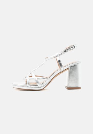 Sølvmetallic sandal med stropper, blokhæl og justerbar ankelspænde, vist i siden mod hvid baggrund.