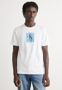 Jaunas vyras dėvi baltą Calvin Klein marškinėlį su mėlynu kvadratiniu logotipu ir šviesiai mėlynas džinsus, stovėdamas prieš neutralų foną.