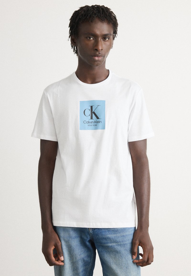 Jaunas vyras dėvi baltą Calvin Klein marškinėlį su mėlynu kvadratiniu logotipu ir šviesiai mėlynas džinsus, stovėdamas prieš neutralų foną.