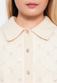 Detailná fotografia osoby v smotanovom pletenom kardigane s perlovo-bielymi gombíkmi a okrúhlym golierom, zdobenom malými striebornými flitrami v kvetinových vzoroch.