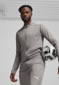 Graue Sportjacke mit Reißverschluss, strukturiertem Stoff und taillierter Passform, kombiniert mit passenden Hosen und einem schwarz-weißen Fußball.