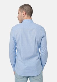 Camisa de manga larga de color azul claro con un sutil patrón repetido, cierre con botones en el frente y cuello estándar. Textura suave y corte ajustado.