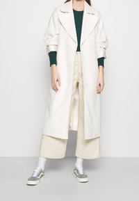 Kvinna som bär en lång vit kappa över en grön topp, krämfärgade vida byxor, vita strumpor och grå sneakers framför en neutral bakgrund.