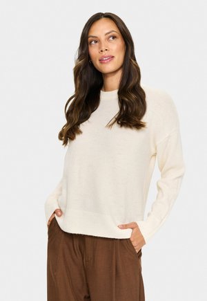 Vrouw met lang golvend haar, gekleed in een off-white trui en bruine broek, met haar handen in haar zakken, die licht omhoog en naar rechts kijkt.