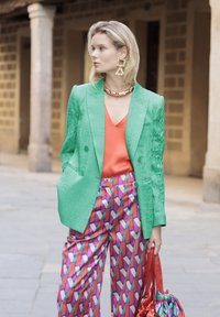 Grön texturerad blazer, orange topp, mönstrade rosa och lila byxor, överdimensionerade örhängen och en glänsande röd väska med rynkade detaljer.