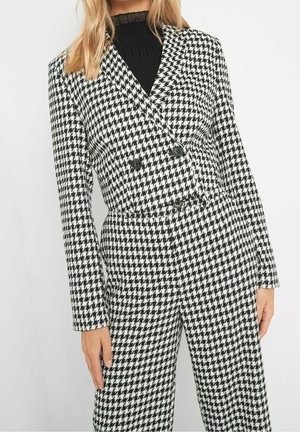 Femme porte un blazer croisé à motif pied-de-poule noir et blanc et un pantalon assorti avec un top noir côtelé en dessous.