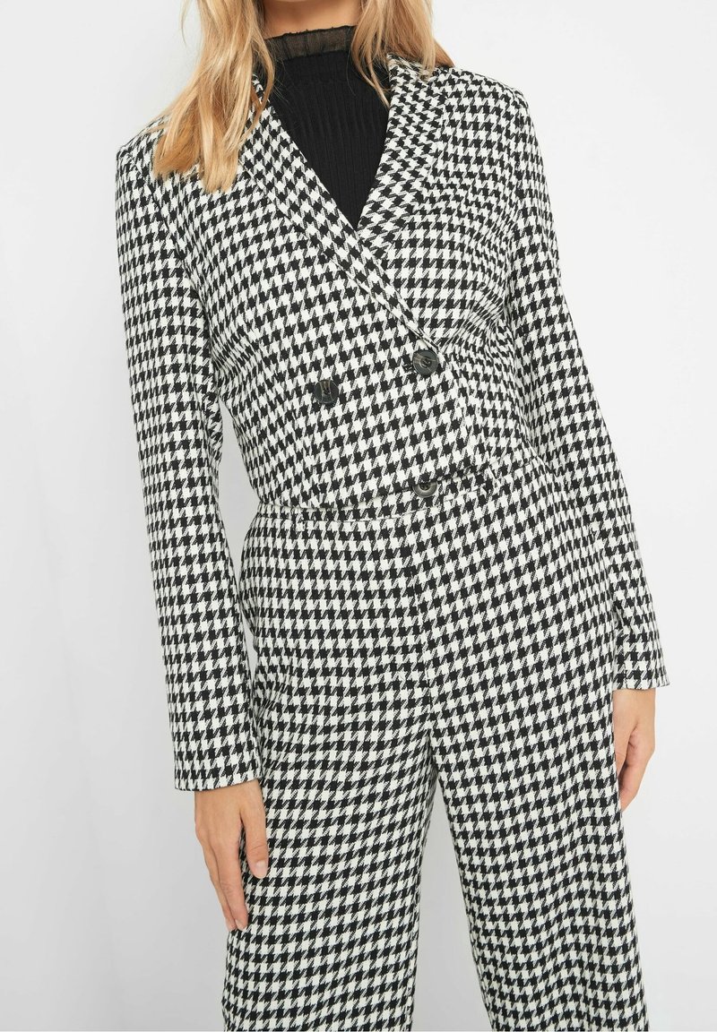 Femme porte un blazer croisé à motif pied-de-poule noir et blanc et un pantalon assorti avec un top noir côtelé en dessous.