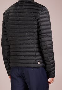 Svart puffjacka med quiltat mönster, med hög krage och en rund logotyp accent på vänster sida nära fållen.