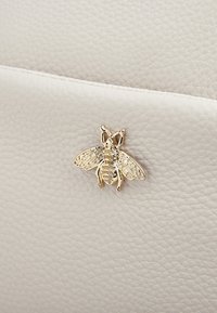 Beżowa skórzana torba o teksturowanej powierzchni, z złotym emblematem pszczoły jako wyrazistym detalem. Prosty design z eleganckim zapięciem.