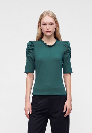 Femme aux cheveux blonds lisses portant un haut vert à manches courtes avec des épaules froncées et un pantalon noir, debout devant un fond uni.