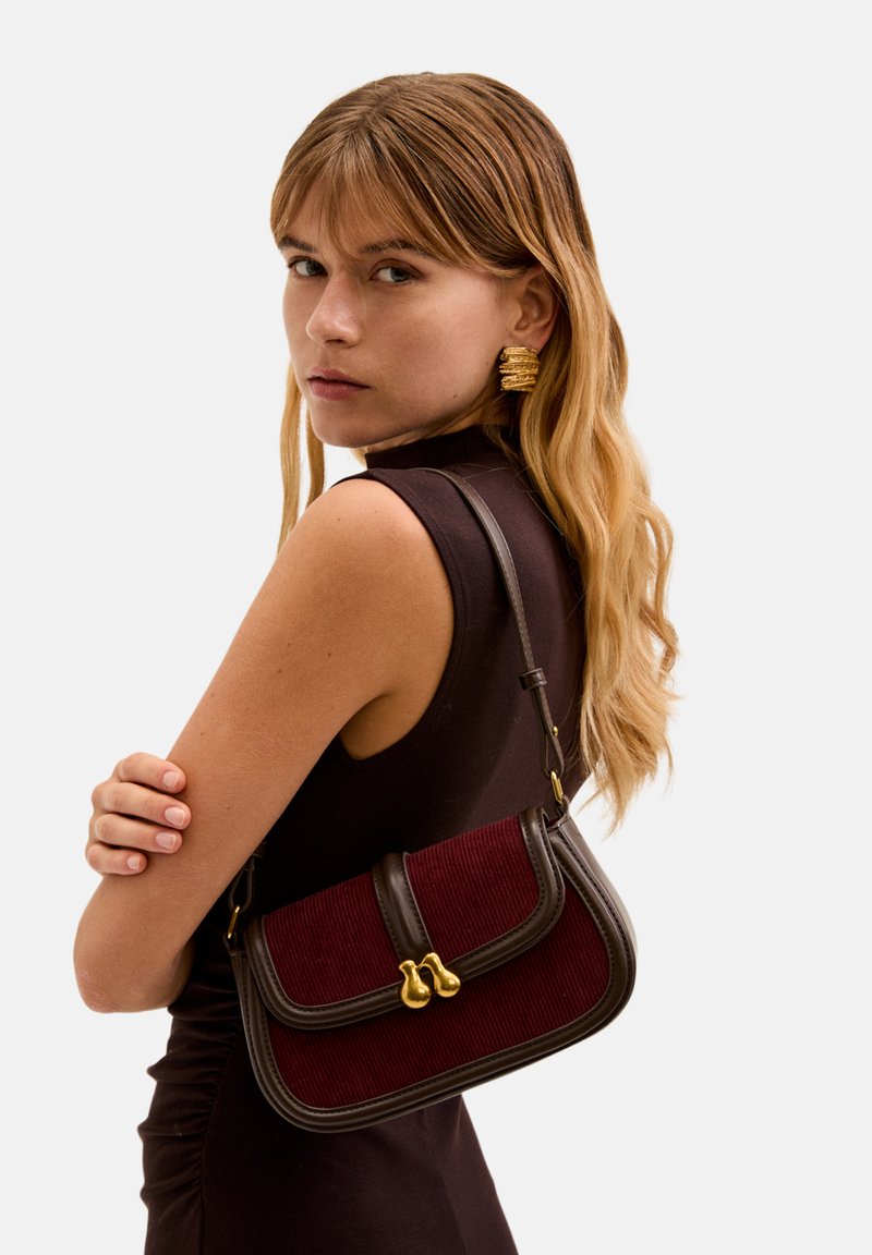 Červená kordurovová kabelka s hnědými koženými akcenty a zlatým zapínáním, s kompaktním obdélníkovým tvarem a krátkým ramenním popruhem.