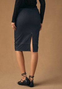 Ciemnoniebieska ołówkowa spódnica dżinsowa z rozcięciem z tyłu, kontrastującym żółtym przeszyciem, noszona z czarnymi płaskimi pantoflami sznurowanymi. Gładka tekstura, długość do połowy łydki.