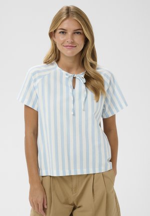 Ung kvinde med langt blondt hår iført en lyseblå og hvid, vertikalt stribet kortærmet top og beige bukser, hænderne i lommerne.