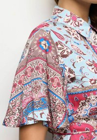 Korte mouw shirt van een lichtblauwe stof met bloemen- en geometrische patronen in bruin, roze en blauw, met een kraag halslijn.