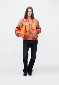 Brązowa kurtka typu bomber z dużymi czerwonymi kwiatowymi wzorami, pomarańczowa kopertówka, ciemne dżinsy i czarne buty. Gładka tkanina i dopasowany krój.