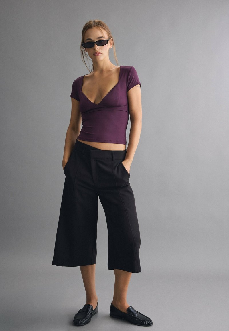 Jeune femme portant un haut violet à col en V, un pantalon noir ample coupé court, des mocassins noirs et des lunettes de soleil noires étroites, debout les mains dans les poches.