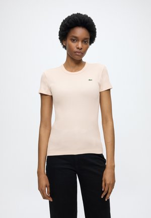 Vrouw met kort krullend haar, die een aansluitend beige T-shirt met korte mouwen en een zwarte broek draagt, staand tegen een effen lichte achtergrond.