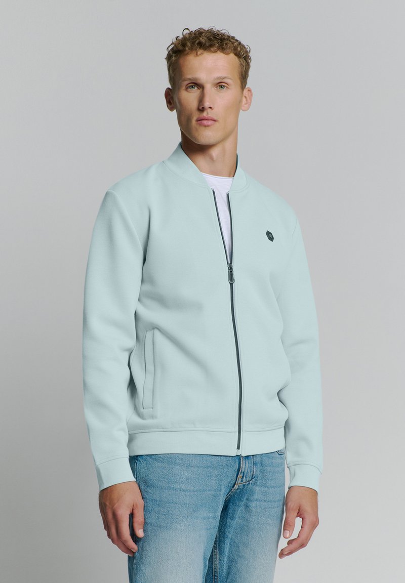 Lichtblauwe bomberjack met ritssluiting en een gestructureerde afwerking, zijzakken en een minimalistisch logo op de linkerborst. Draagt over een wit shirt.