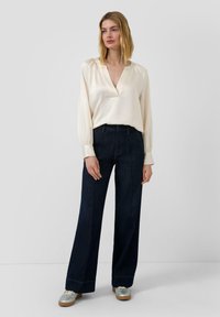 Kremowa bluzka z głębokim dekoltem w kształcie litery V i marszczonymi ramionami, w połączeniu z dżinsami o wysokim stanie i szerokich nogawkach w ciemnym kolorze. Srebrne buty.