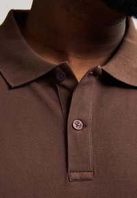 Brązowa koszulka polo z teksturowanej tkaniny, z złożonym kołnierzykiem i dwoma ciemnymi guzikami na listwie. Minimalne widoczne szwy.