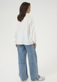 White long-sleeve shirt with gathered shoulders, paired with wide-leg blue denim jeans featuring contrasting seams. Casual footwear visible.